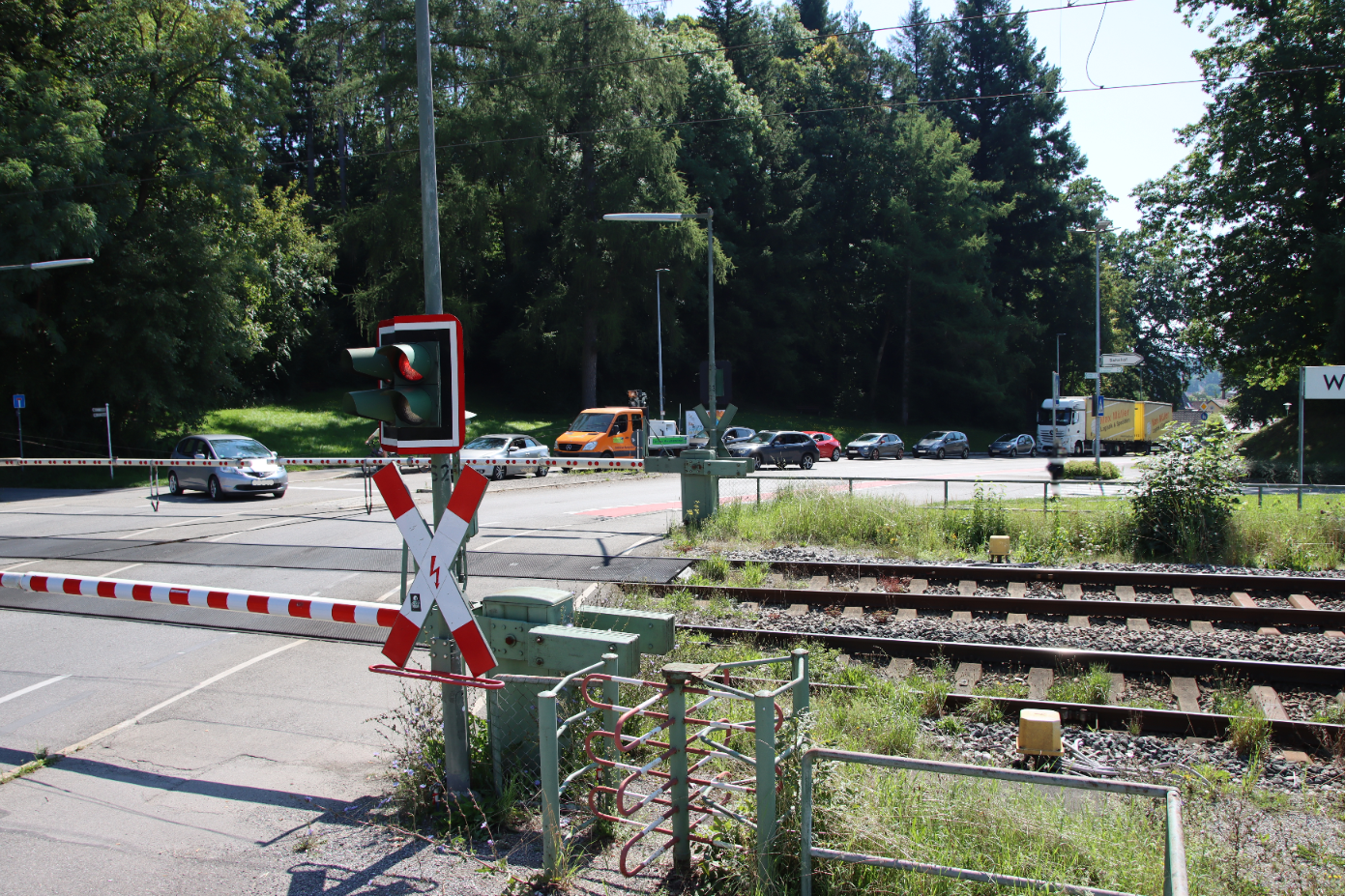 Das Bild zeit einen Bahnübergang mit heruntergelassenen Schranken und eine rote Ampel. Mehrere wartende Fahrzeuge