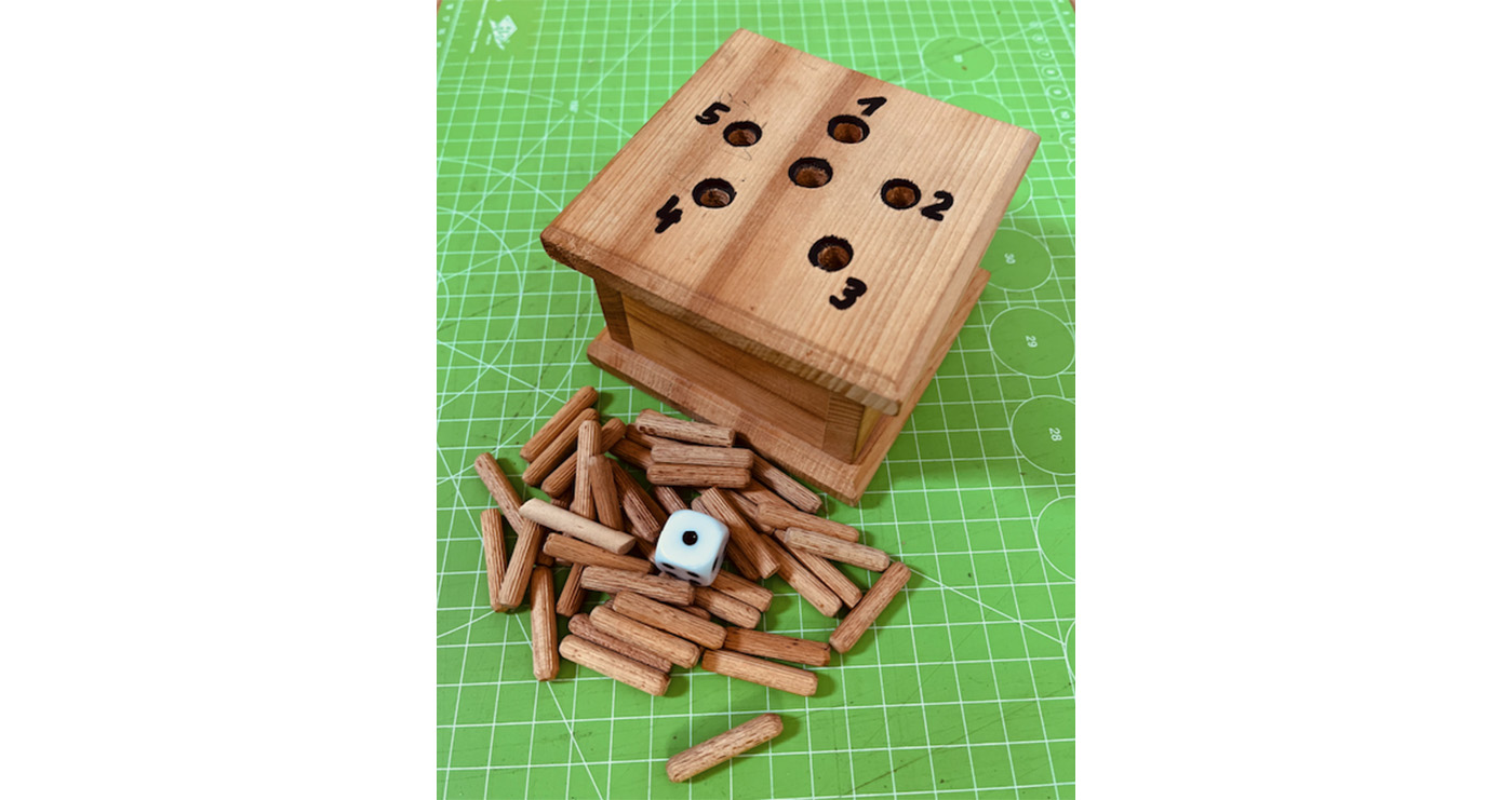Das Foto zeigt das Würfelspiel "Ab in die Box". Viereckige Box aus Holz mit 6 Löchern, mehrere kurze Holzstifte und ein Würfel