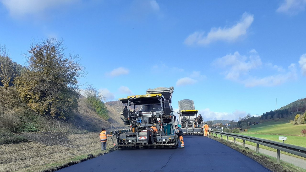 Man sieht Baustellenfahrzeug das neuen Asphalt auf einer Straße walzt