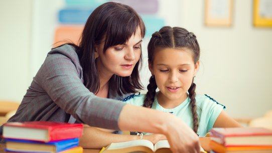 Junge Lehrerin mit Schülerin an einem Schreibtisch mit vielen Büchern darauf