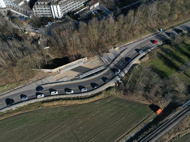 Luftbild einer Straßenbaustelle. Die Straße führt parallel zu einer Brücke über eine Behelfsumfahrung. Die Brücke ist fast fertiggstellt, in der Mitte hat sie bereits eine Schicht Asphalt, an den Übergängen zur Straße liegt der Kies frei. 