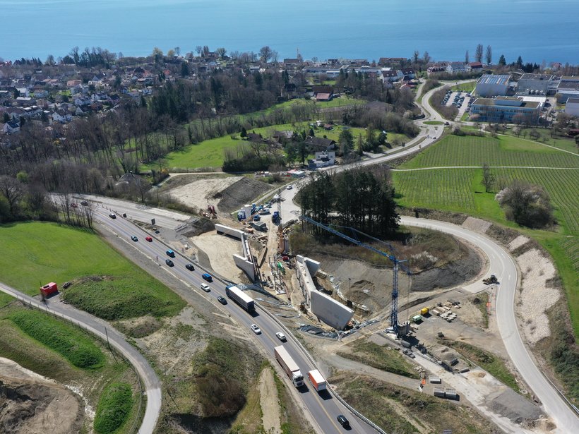 Ein Luftbild einer Baustelle, im Hintergrund der Bodensee. Auf der Baustelle sind zwei große Betonwände sichtbar, zischen denen Kräne ein Stahlgerüst aufbauen.