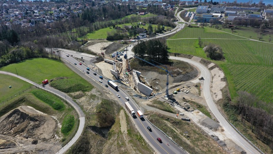 März 2024: Die Widerlager sind fertig betoniert, jetzt beginnt der Aufbau des Traggerüsts, auf dem später die Brücke betoniert wird. Ein Luftbild einer Baustelle, im Hintergrund der Bodensee. Auf der Baustelle sind zwei große Betonwände sichtbar, zischen denen Kräne ein Stahlgerüst aufbauen.