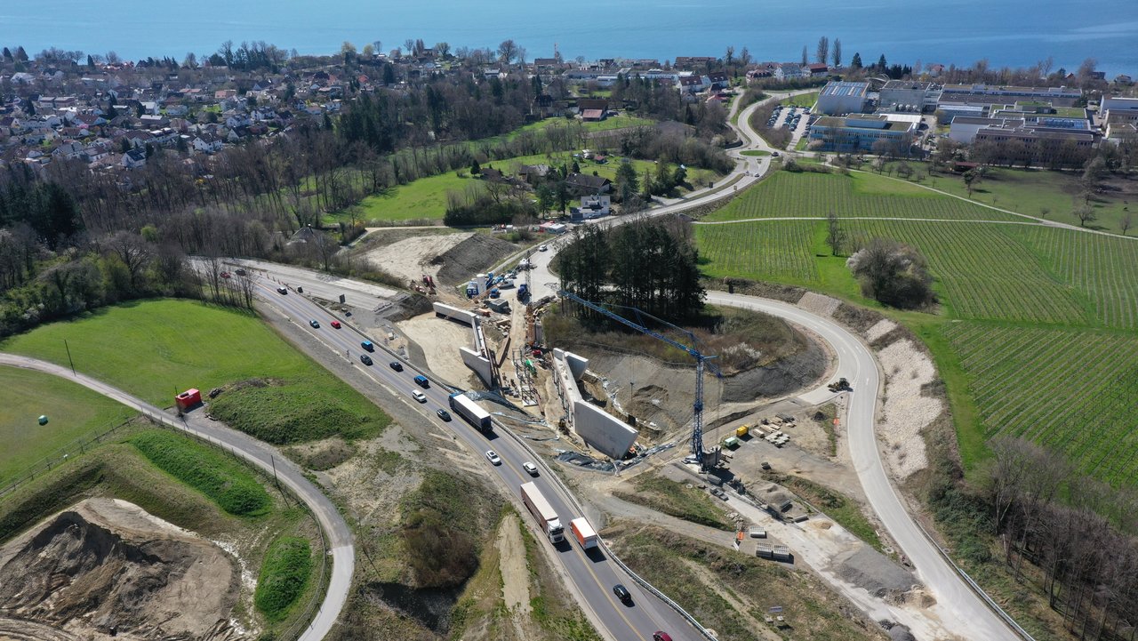 Ein Luftbild einer Baustelle, im Hintergrund der Bodensee. Auf der Baustelle sind zwei große Betonwände sichtbar, zischen denen Kräne ein Stahlgerüst aufbauen.