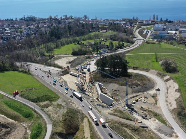 März 2024: Die Widerlager sind fertig betoniert, jetzt beginnt der Aufbau des Traggerüsts, auf dem später die Brücke betoniert wird. Ein Luftbild einer Baustelle, im Hintergrund der Bodensee. Auf der Baustelle sind zwei große Betonwände sichtbar, zischen denen Kräne ein Stahlgerüst aufbauen.