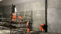 Vier Arbeiter betonieren eine Wand in einem Tunnel. Zwei Arbeiter stehen auf einem rollbaren Gerüst, zwei am Boden. Auf einem Teil der Wand glänzt der Beton, weil er noch feucht ist. 