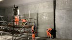 Vier Arbeiter betonieren eine Wand in einem Tunnel. Zwei Arbeiter stehen auf einem rollbaren Gerüst, zwei am Boden. Auf einem Teil der Wand glänzt der Beton, weil er noch feucht ist. 