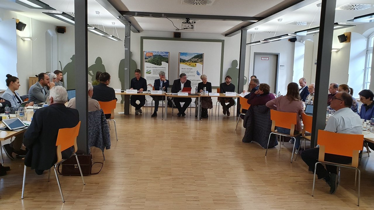 Das Foto zeigt eine Sitzung in einem großen Raum der mehrere Personen, mit Blick auf einen Tisch an dem die Redner sitzen, beiwohnen