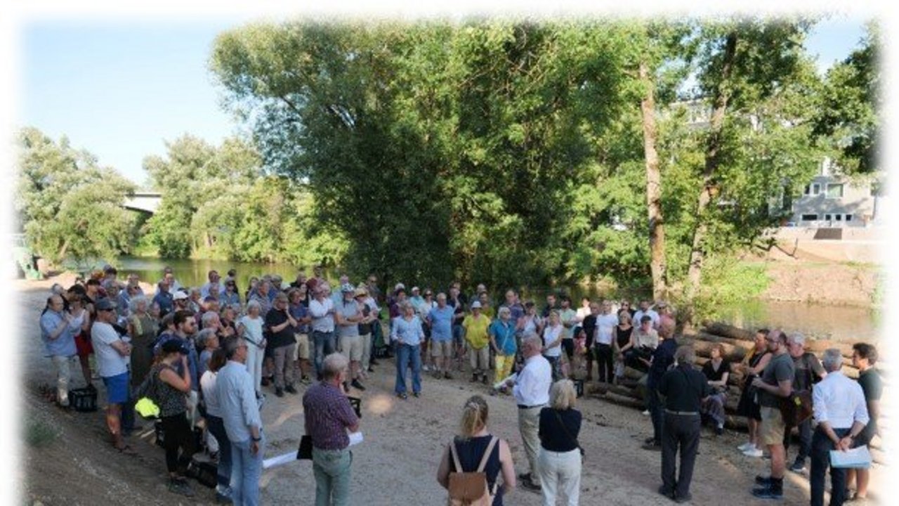 Eine Gruppe von Menschen - rund 150 Personen stehen auf einer Baustelle am Neckarufer und hören einem Vortragenden zu.