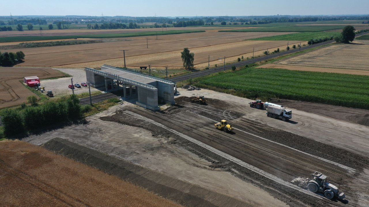 Man sieht die Baustelle mit Baustellenfahrzeugen, eine Brücke über die Bahngleise im Bau, Äcker, Felder von oben und im Hintergrund eine Ortschaft