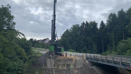 Juli 2025: Die Bohrpfähle für die Widerlager der neuen Brücke werden hergestellt. Ein Bohrgerät neben einer Brücke