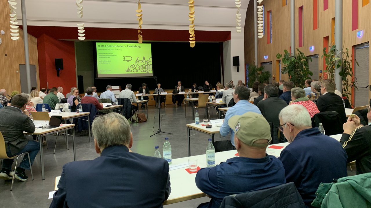 Das Foto zeigt eine Informationsveranstaltung. Mehrere Personen an Tischen in einem großen Raum. Man blickt auf eine Leinwand mit einer Präsentation zum Thema "B 30, Friedrichshafen - Ravensburg"