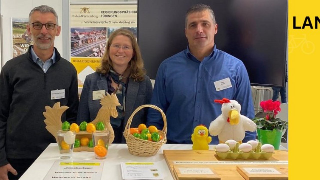Blick in eine Ausstellungshalle des Landwirtschaftlichen Hautfestes. Zwei Kollegen aus der Marktüberwachung Eiermarkt stehen hinter zwei Tischen und lächeln in die Kamera. auf dem Tisch liegen Materialien wie Eierkartons, Gläser mit Wasser und Infoflyer.
