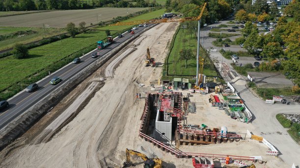 Luftbild einer Straßenbaustelle. Von Links nach rechts: Straße, Abschnitt auf dem Erdarbeiten für eine neue Trasse stattfinden, Baustelle für eine Betonkonstruktion, die tief in den Boden ragt. 