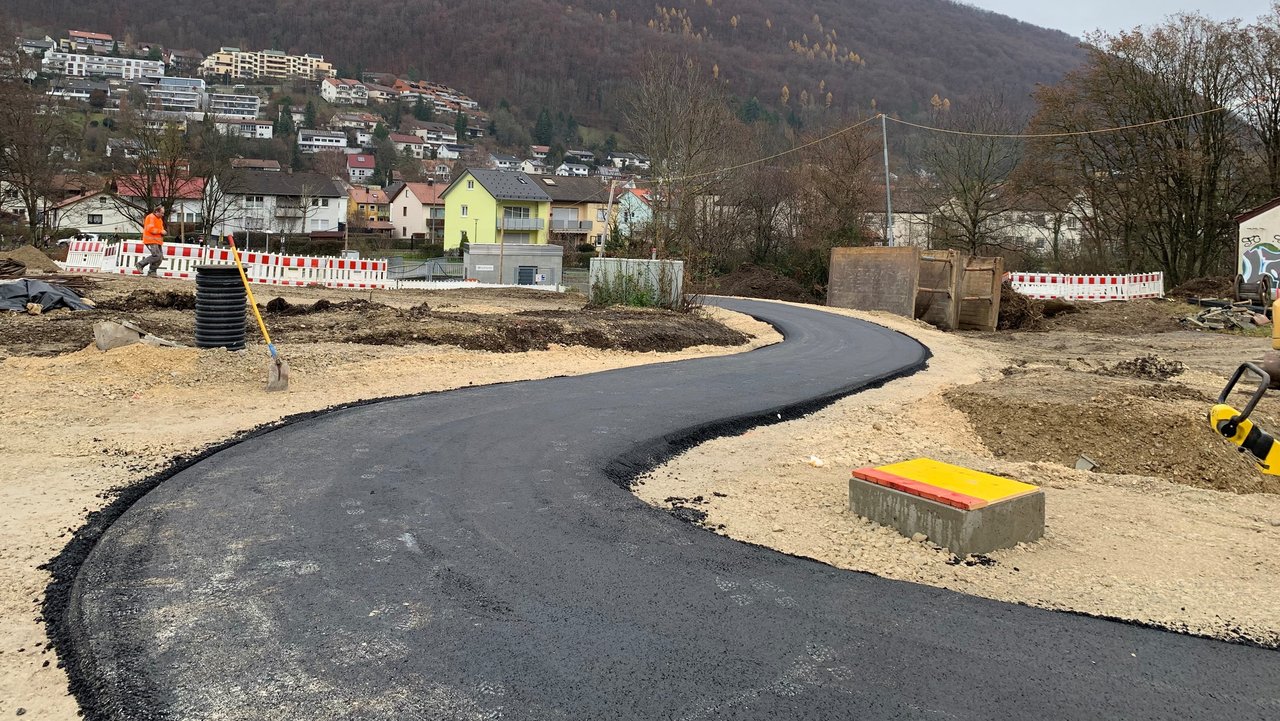 Das Foto zeigt einen frisch geteerten Weg, der auf eine Baustellenabsperrung zuläuft. Im Hintergrund am Hang sieht man eine Ortschaft.