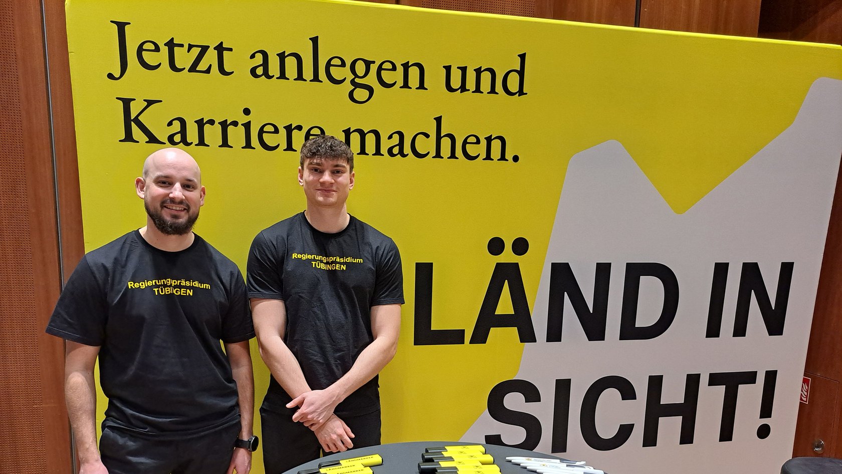 Zwei junge Männer stehen auf einer Ausstellungswand und lächeln.