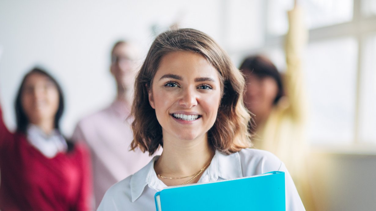 Lehrerin hält eine Mappe und lacht in die Kamera. Im Hintergrund sind 3 weitere LehrerInnen zu sehen, die die Hände hebenn