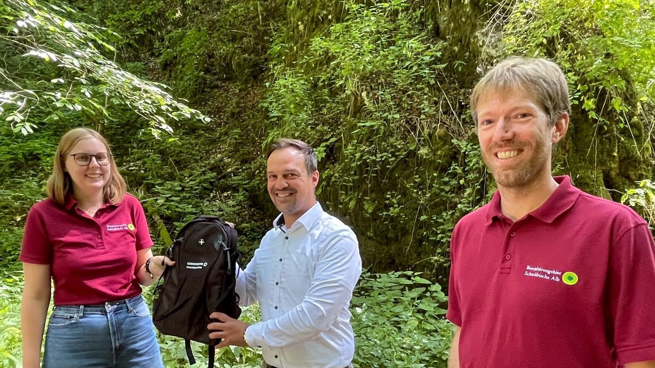 Das Bild zeigt, wie Patrick Eschle Nadine Pluquette einen schwarzen Rucksack überreicht; daneben steht Volker Häring 