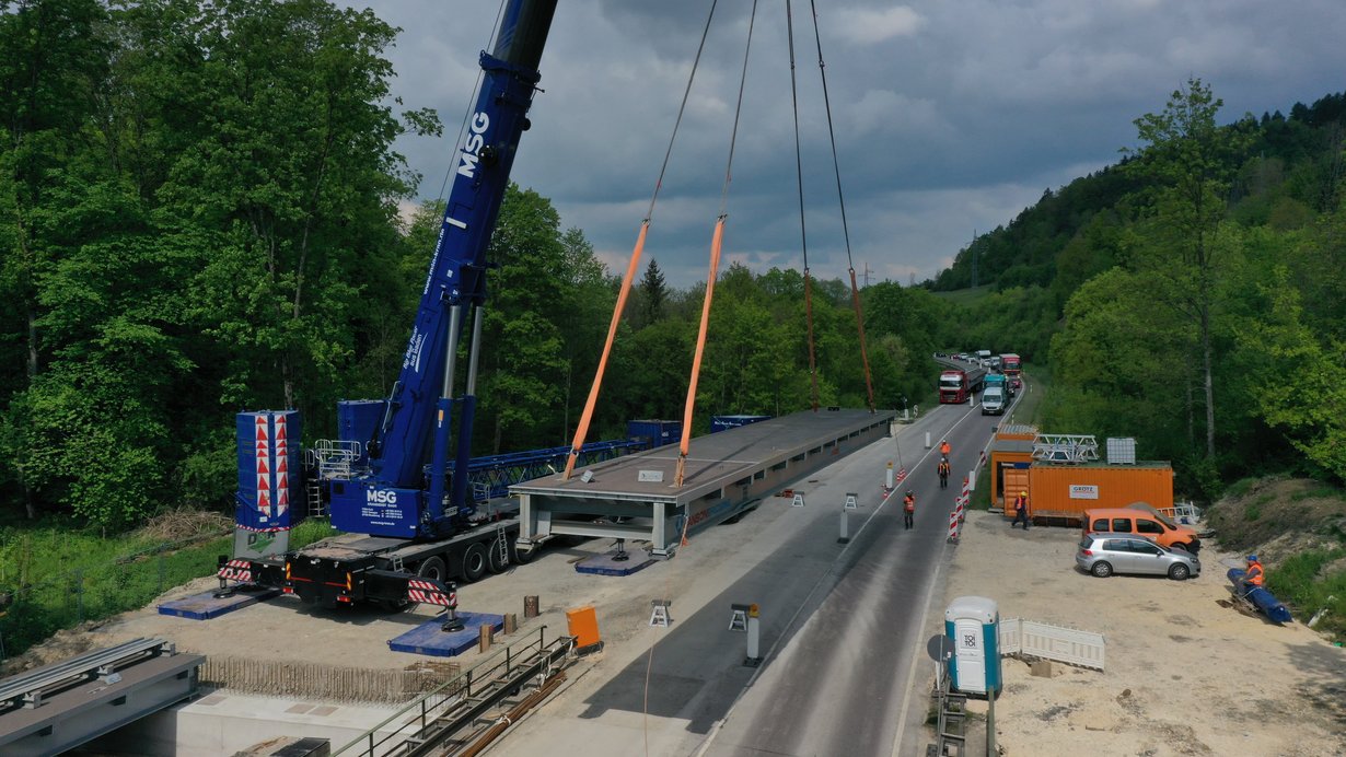 Ein Kran steht neben einer Straße. Er Hebt ein Brückenteil, das direkt neben der Straße liegt, auf circa einen Meter Höhe. Vor dem Kran befinden sich Brücken-Widerlager, auf denen bereits ein Brückenteil abgelegt ist.