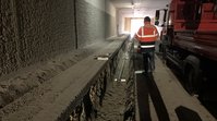 In einem Tunnel läuft ein Graben, in dem eine Röhre liegt. Neben dem Graben steht ein Mann in einer orangenen Bau-Jacke.