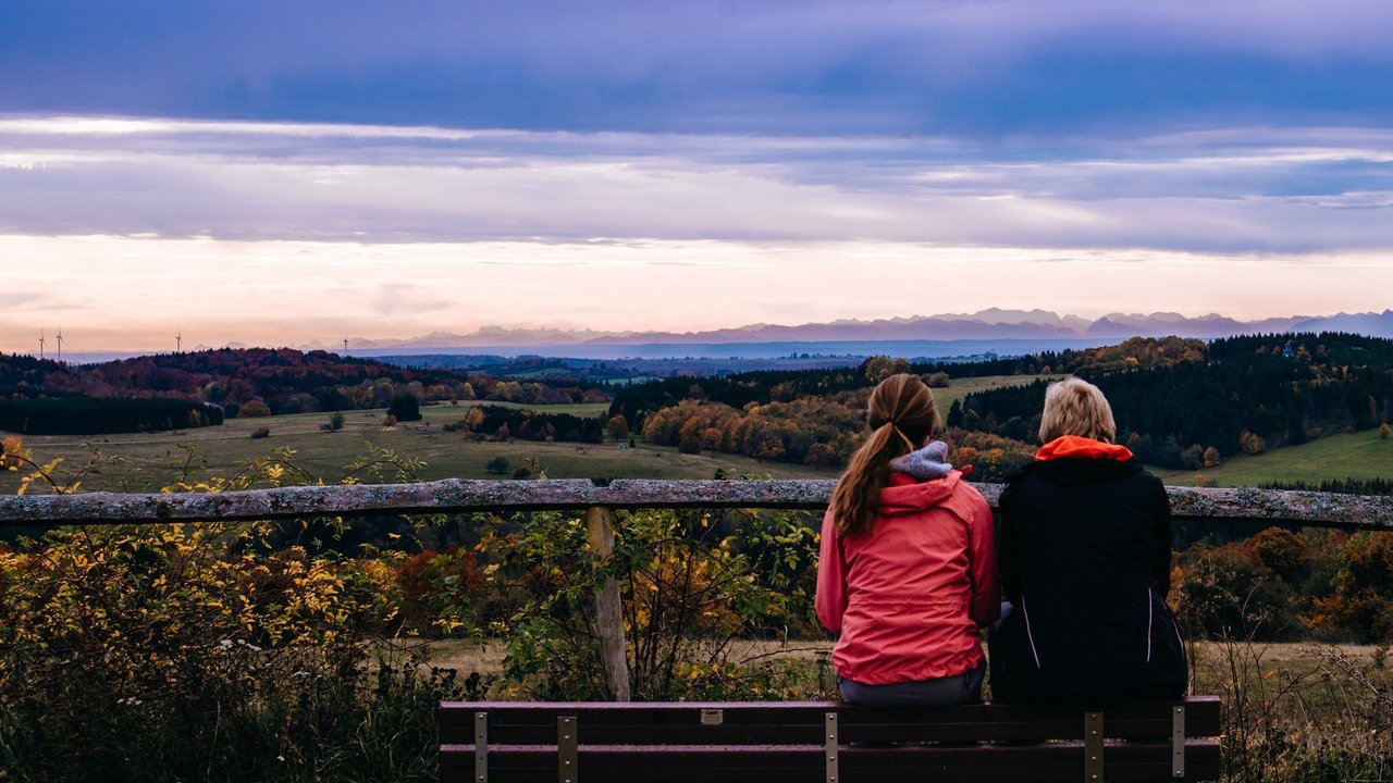 Zwei Frauen sitzen bei Sonnenaufgang auf einem Berg auf einer Bank und schauen ins Tal