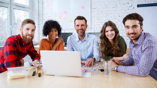 Lehrerinnen und Lehrer sitzen am Tisch und lachen in die Kamera