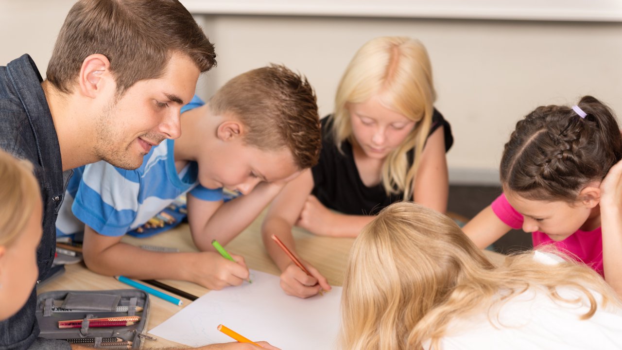 Ein junger Lehrer sitzt mit einer Gruppe aus fünf Schülerinnen und Schülern am Tisch und arbeiten zusammen. 
