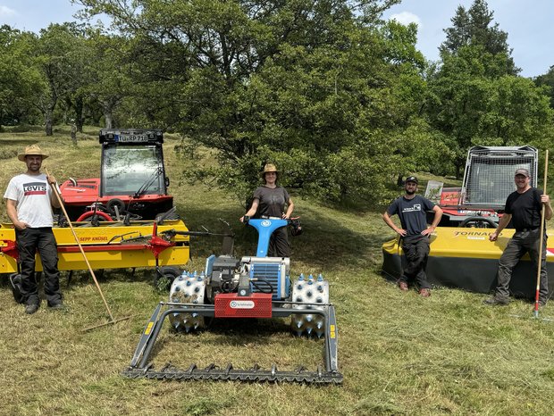 Landschaftspflegetrupp Fünf Mitarbeitende (5 Männer, eine Frau) des Landschaftspflegetrupps lehnen an ihren Arbeitsmaschinen (Traktor, Mäher) und lächeln in die Kamera.