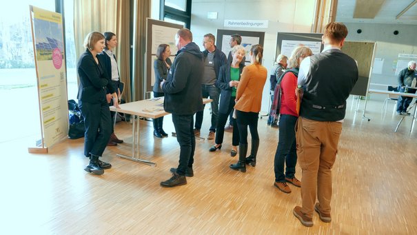 Man sieht einen Tisch mit Materialien, um den herum Menschen stehen und diskutieren. Im Hintergrund sind Stellwände mit Plakaten zu sehen.