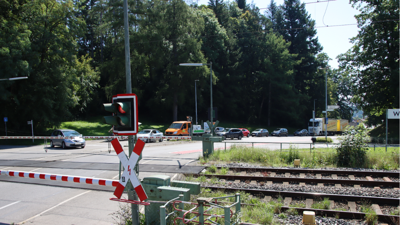 Das Bild zeit einen Bahnübergang mit heruntergelassenen Schranken und eine rote Ampel. Mehrere wartende Fahrzeuge