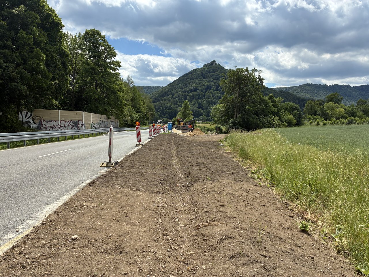 Das Foto zeigt eine Straße die die durch mehrere Stehlen von einem unbefestigten Streifen abgegrenzt wird. Im Hintergrund sieht man einen Burg auf einem Berg.