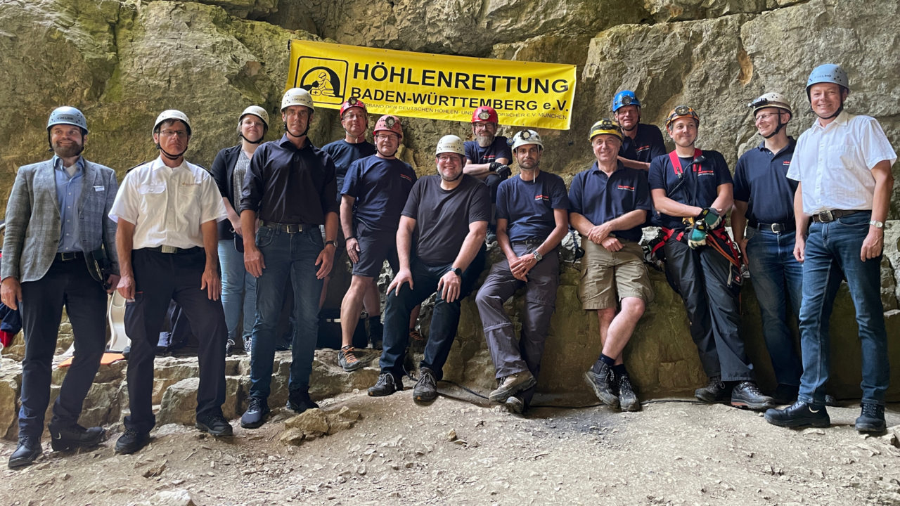 Das Gruppenfoto im Eingangsbereich der Falkensteiner Höhle zeigt Mitglieder der Höhlenrettung Baden-Württemberg e.V.. Auf der rechten Seite steht Herr Regierungspräsident Klaus Tappeser. Im oberen Bildbereich ist ein Banner mit dem Logo des Vereins an der Höhlenwand zu sehen.