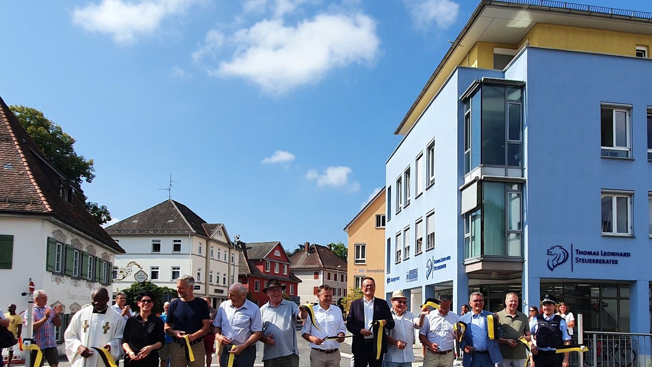 Das Foto zeigt den Banddurchschnitt bei der Verkehrsfreigabe der Eschachbrücke in Leutkirch. 12 Personen durchschneiden das Band. Im Hintergrund Gebäude in Leutkirch
