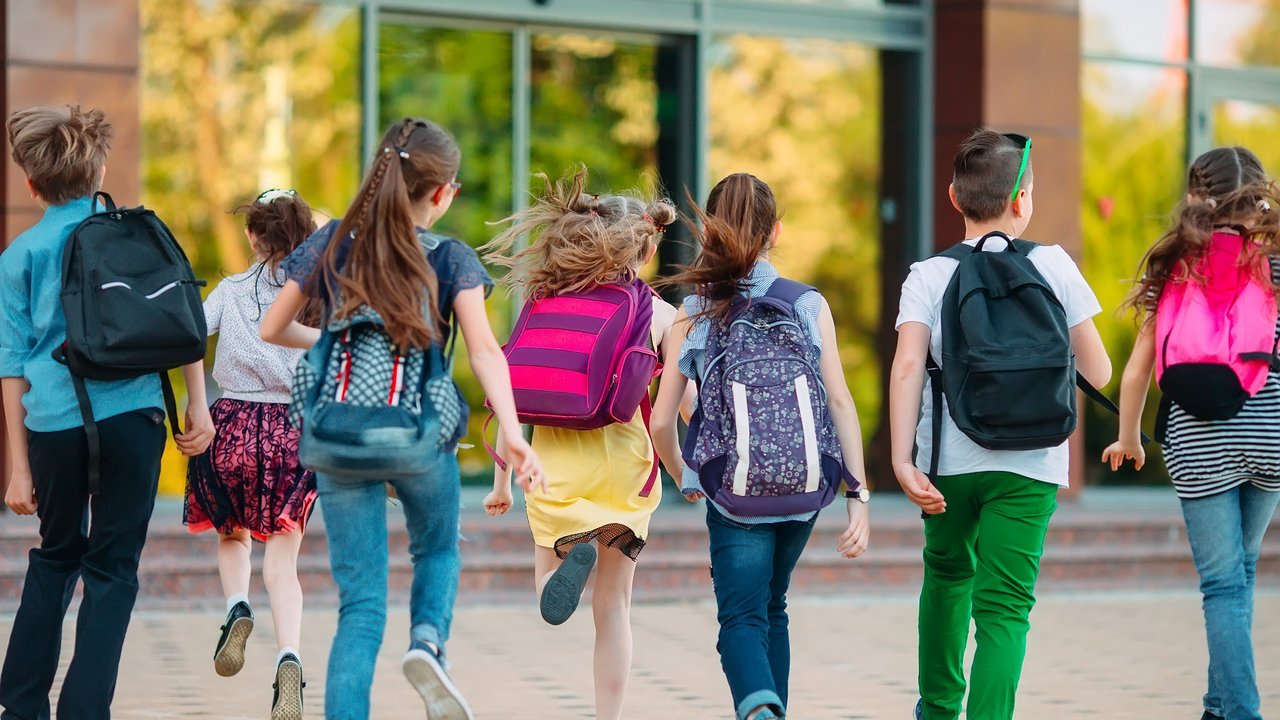 7 Schulkinder mit Rucksäcken rennen auf ein Schulgebäude zu
