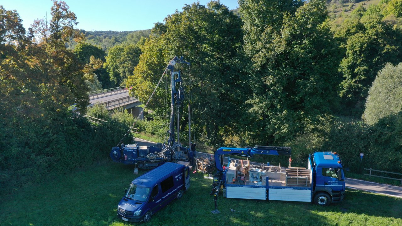 Das Bild zeigt eine Baugrunduntersuchung auf einer Wiese mit Bäumen, einer Straße und einer Brücke im Hintergrund; mehrere Baustellenfahrzeuge