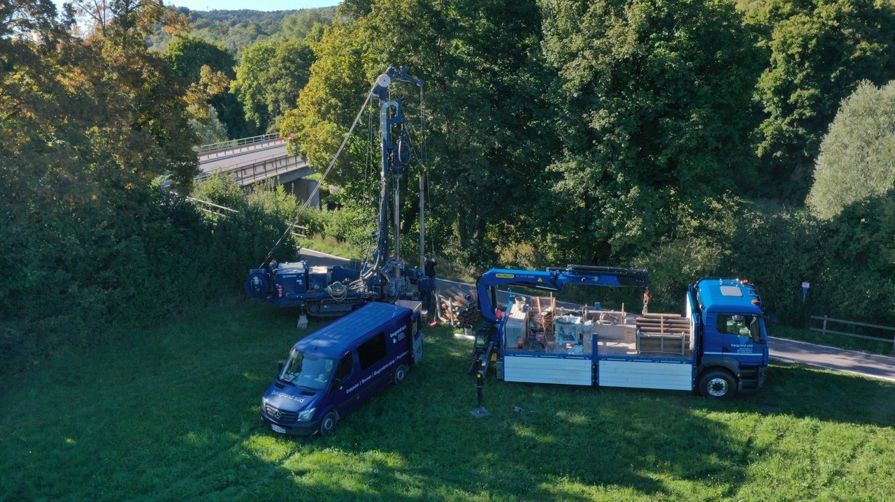 Das Bild zeigt eine Baugrunduntersuchung auf einer Wiese mit Bäumen, einer Straße und einer Brücke im Hintergrund; mehrere Baustellenfahrzeuge