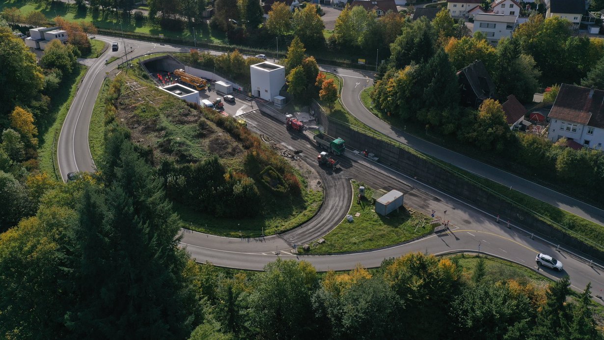 Luftbild einer Baustelle vor einem Tunnelportal, auf der sich mehrere Baumaschinen und LKW befinden.