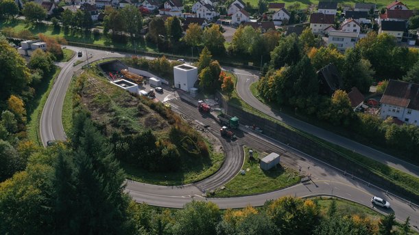 Luftbild einer Baustelle vor einem Tunnelportal, auf der sich mehrere Baumaschinen und LKW befinden.