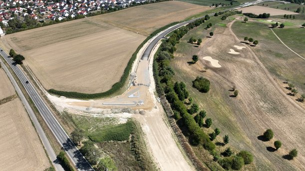 Luftbild einer Straßenbaustelle. Zwei noch nicht asphaltierte Äste einer neuen Straße schließen an eine bestehende an. 