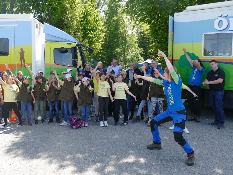 Vor den beiden Ökomobilen steht eine Gruppe Kinder und die Ökomobilistin Sabine Reußink, welche die Gruppe zu einem Bewegungsspiel anleitet.