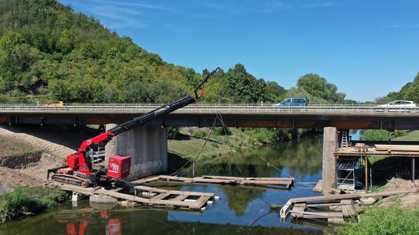 Foto einer Straßenbrücke über einen Fluss von der Seite. Unter der Fahrbahn verlaufen große Stahlträger, auf einer Plattform im Fluss steht eine Art Kran. 