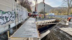 Ein LKW mit einem großen Ausleger steht neben einer Baustelle. Aus dem Ausleger des LKW fließt Beton in eine Schalnug, den Menschen mit Schaufeln verteilen.