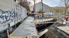 Ein LKW mit einem großen Ausleger steht neben einer Baustelle. Aus dem Ausleger des LKW fließt Beton in eine Schalnug, den Menschen mit Schaufeln verteilen.