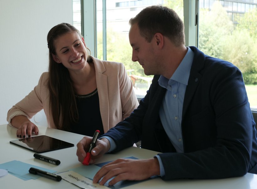 Eine junge Frau und ein junger Mann sitzen am Tisch, die Frau lächelt den Kollegen an und zeigt auf ein Tablet.