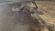 Ein breiter und tiefer Graben, der nach oben breiter wird. In der Sohle des Grabens ist ein Kipper-LKW zu sehen. Ein Bagger steht auf einer Rampe auf ungefähr halber Höhe des Grabens und schaufelt Erde. 