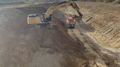Ein breiter und tiefer Graben, der nach oben breiter wird. In der Sohle des Grabens ist ein Kipper-LKW zu sehen. Ein Bagger steht auf einer Rampe auf ungefähr halber Höhe des Grabens und schaufelt Erde. 