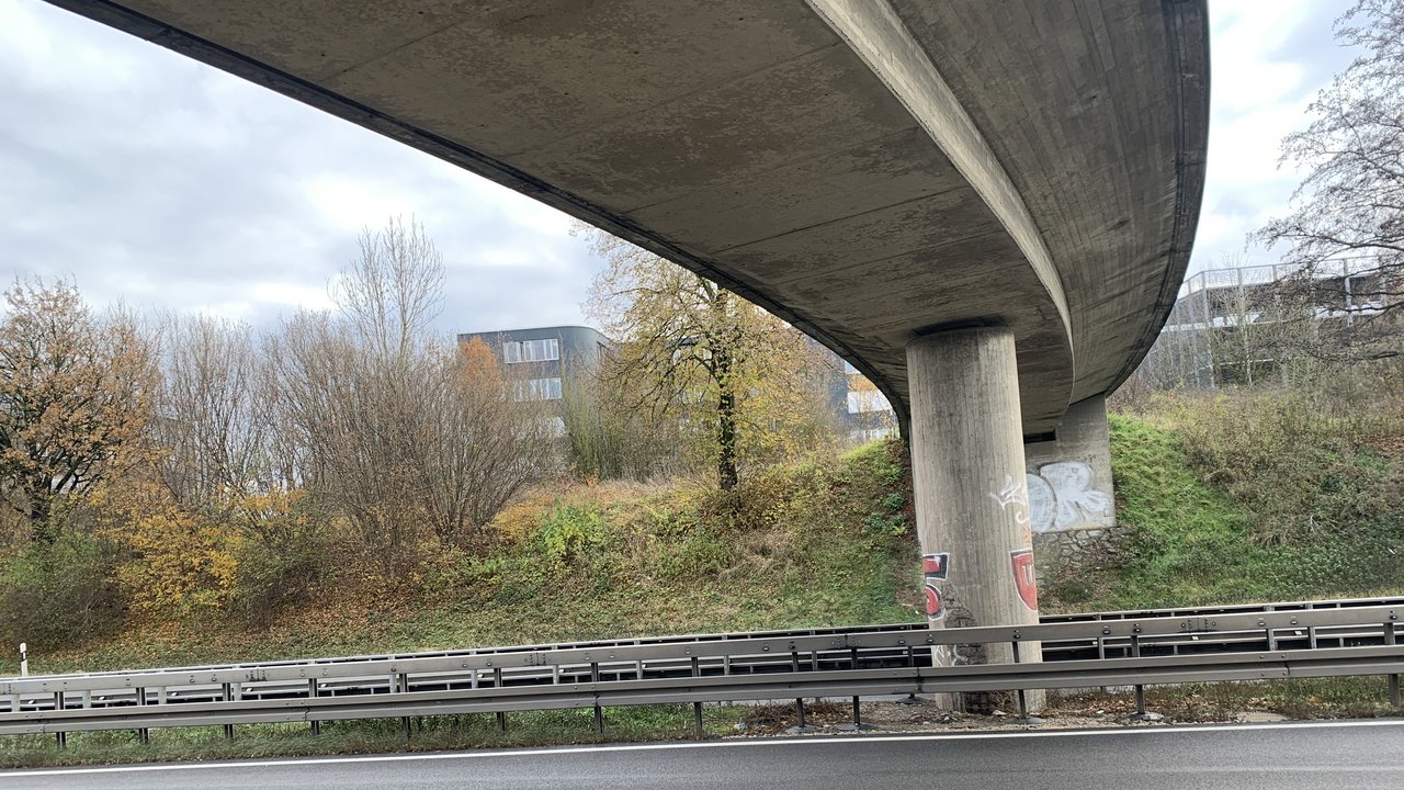 Das Foto zeigt eine Überführung über die B 28. Am Rand der Straße sieht man Leitplanken und einen kleiner mit Sträuchern bewachsener Hang. Im Hintergrund sieht man Wohnblöcke.