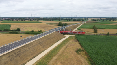 Luftbild einer Straßenbrücke, die über die Gleise führt. Auf den Gleisen fährt ein roter Regionalzug. 