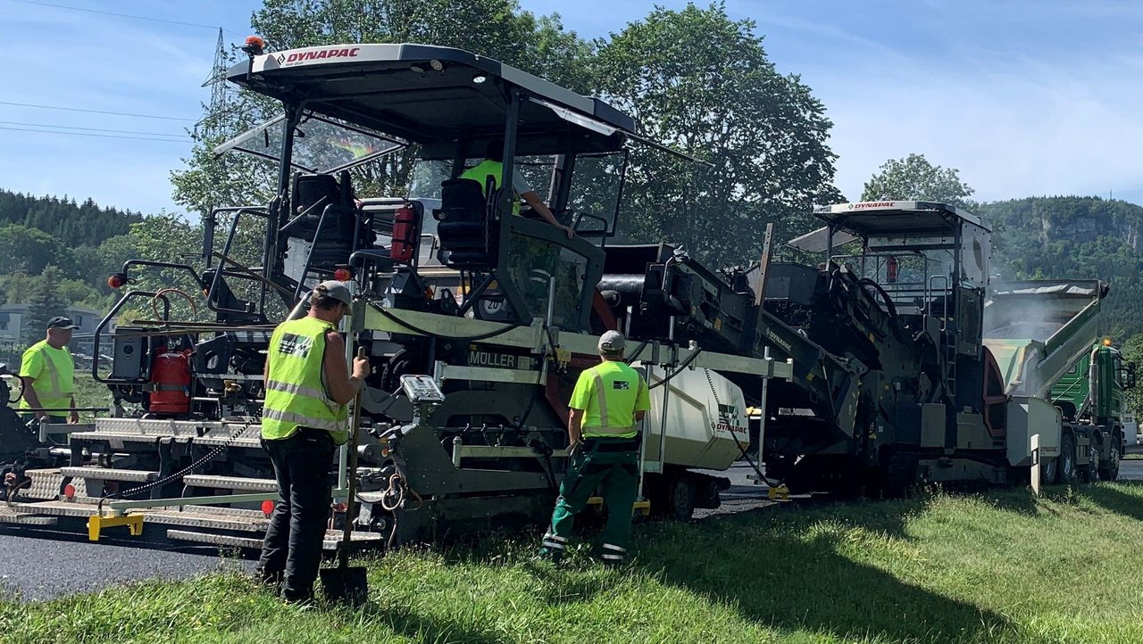 Man sieht ein großes Baustellenfahrzeug bei Asphaltierarbeiten und vier Personen in gelben Warnwesten