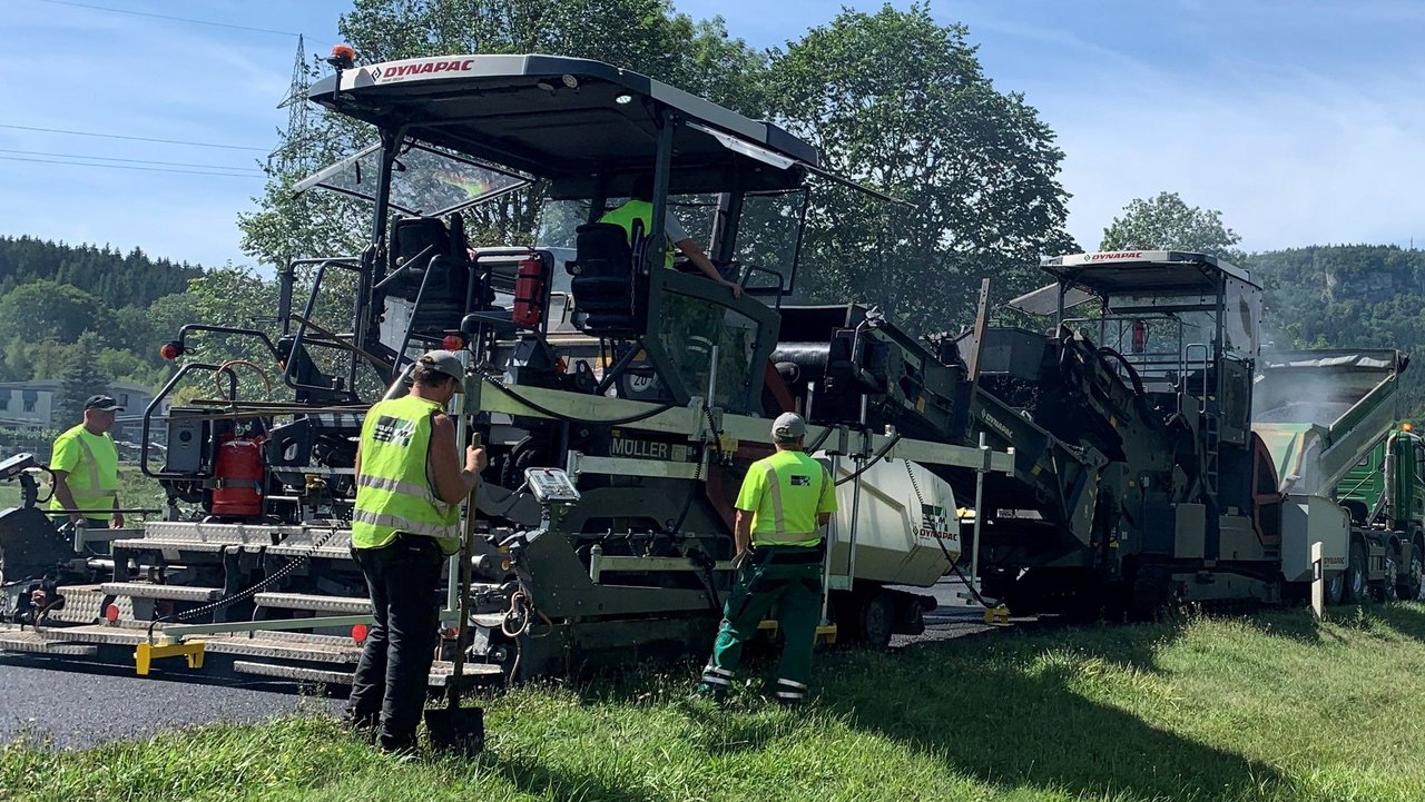 Man sieht ein großes Baustellenfahrzeug bei Asphaltierarbeiten und vier Personen in gelben Warnwesten